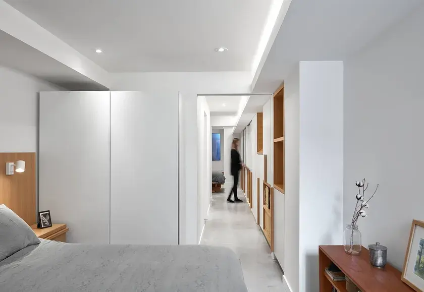 two bedrooms connected by a long narrow hallway with one long continuous millwork wall and a tall woman at the end. The wall contains white cabinets and pocket doors