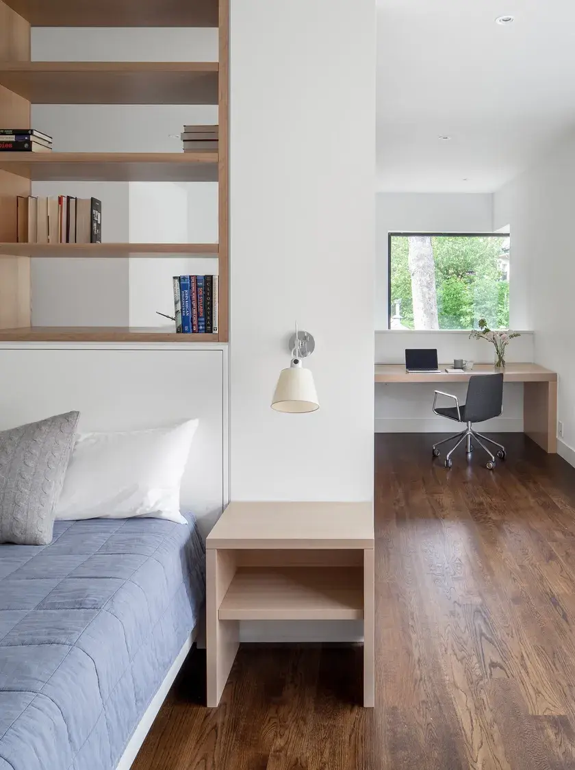 close up of a bed and nightstand in the foreground against a dividing wall with a custom millwork bookshelf and an office desk in the background